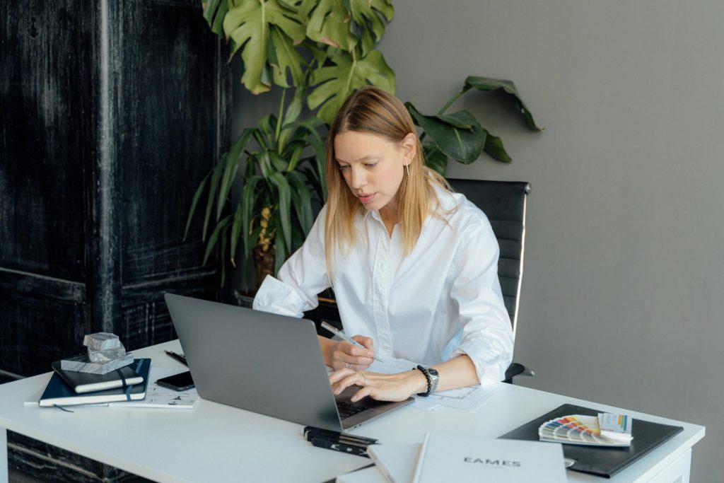 Femme concentrée en chemise blanche travaillant sur un ordinateur portable au bureau avec des plantes et des livres.