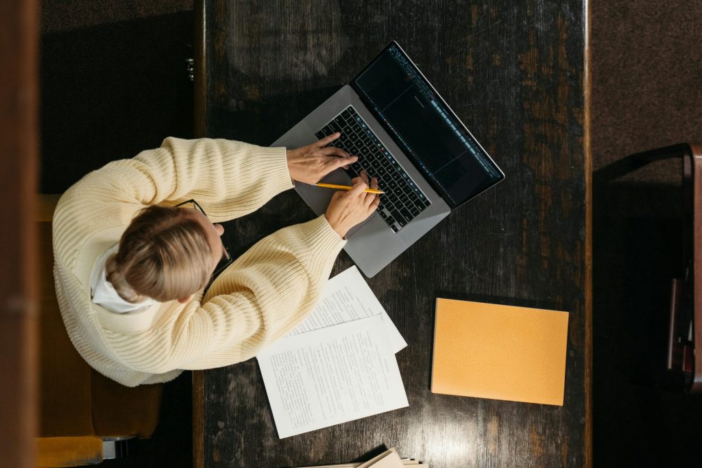 Personne dans un pull confortable travaillant sur un ordinateur portable avec des documents sur un bureau en bois, vue d'en haut.